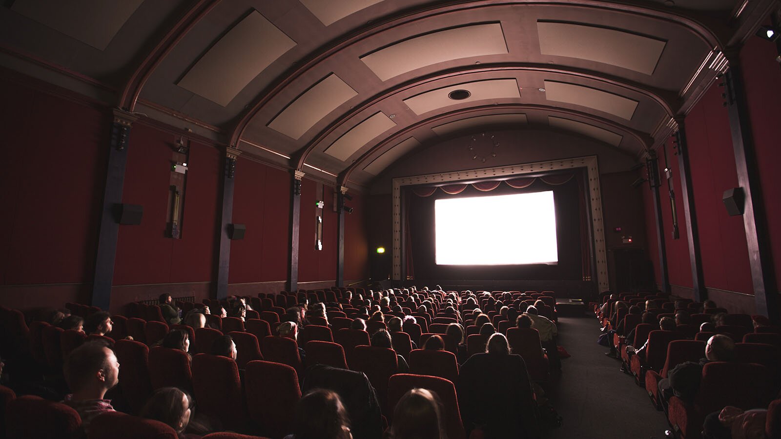 Zdjęcie przedstawia wnętrze starej sali kinowej. Ujęcie jest wykonane z tylnej części sali. Widoczne są rzędy bordowych siedzeń, na których siedzą ludzi. W głębi widoczny jest biały, prostokątny ekran osadzony w ramie. Pomieszczenie ma ściany w kolorze bordowym, podzielone na segmenty za pomocą pionowych półkolumn, oraz łukowate sklepienie z drewnianymi, ciemnobrązowymi kasetonami.