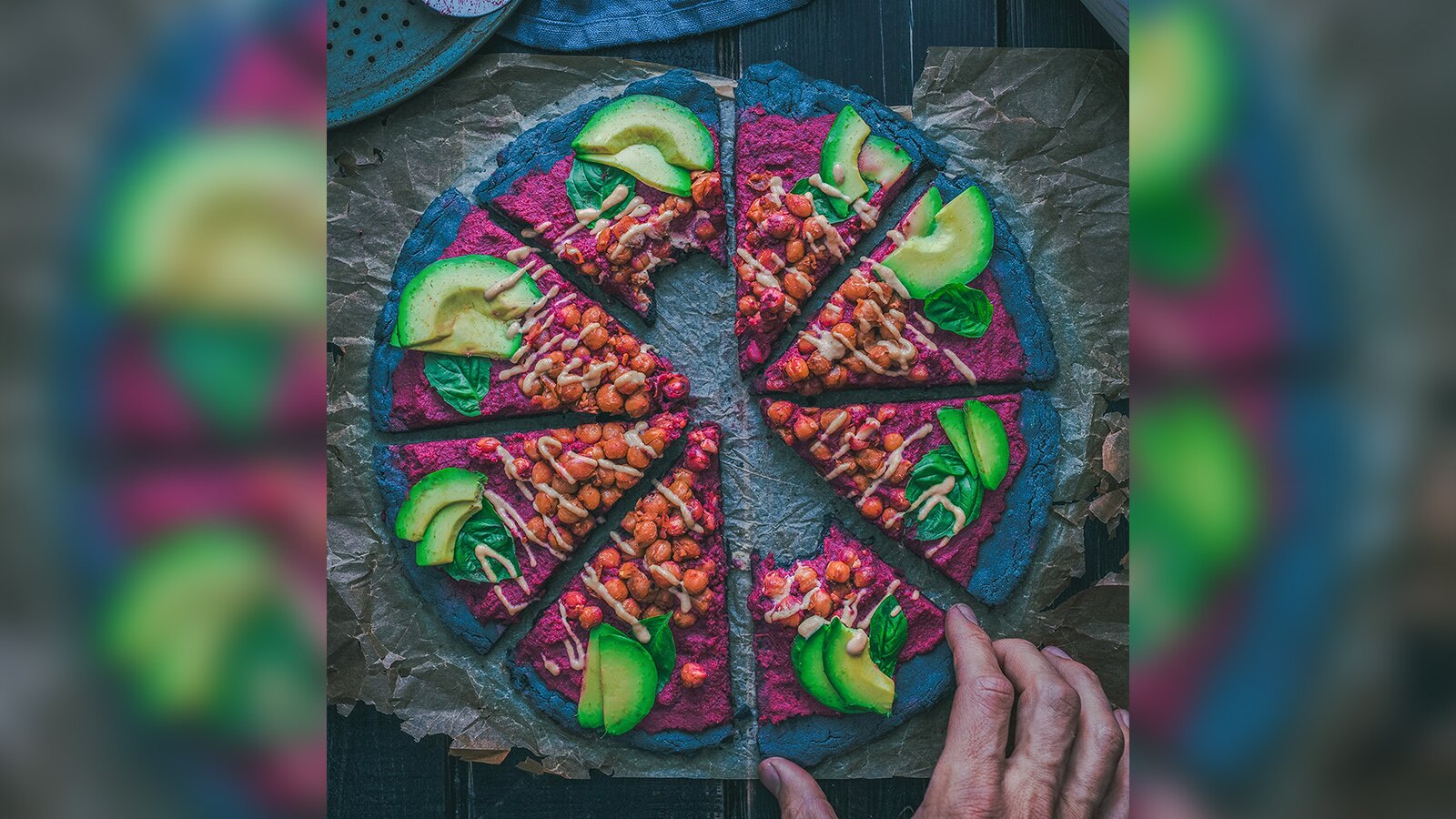 Zdjęcie przedstawia wegańską pizzę. Ciasto jest czarne, a na nim rozsmarowano bordowy sos, na którym ułożono ciecierzycę oraz awokado.