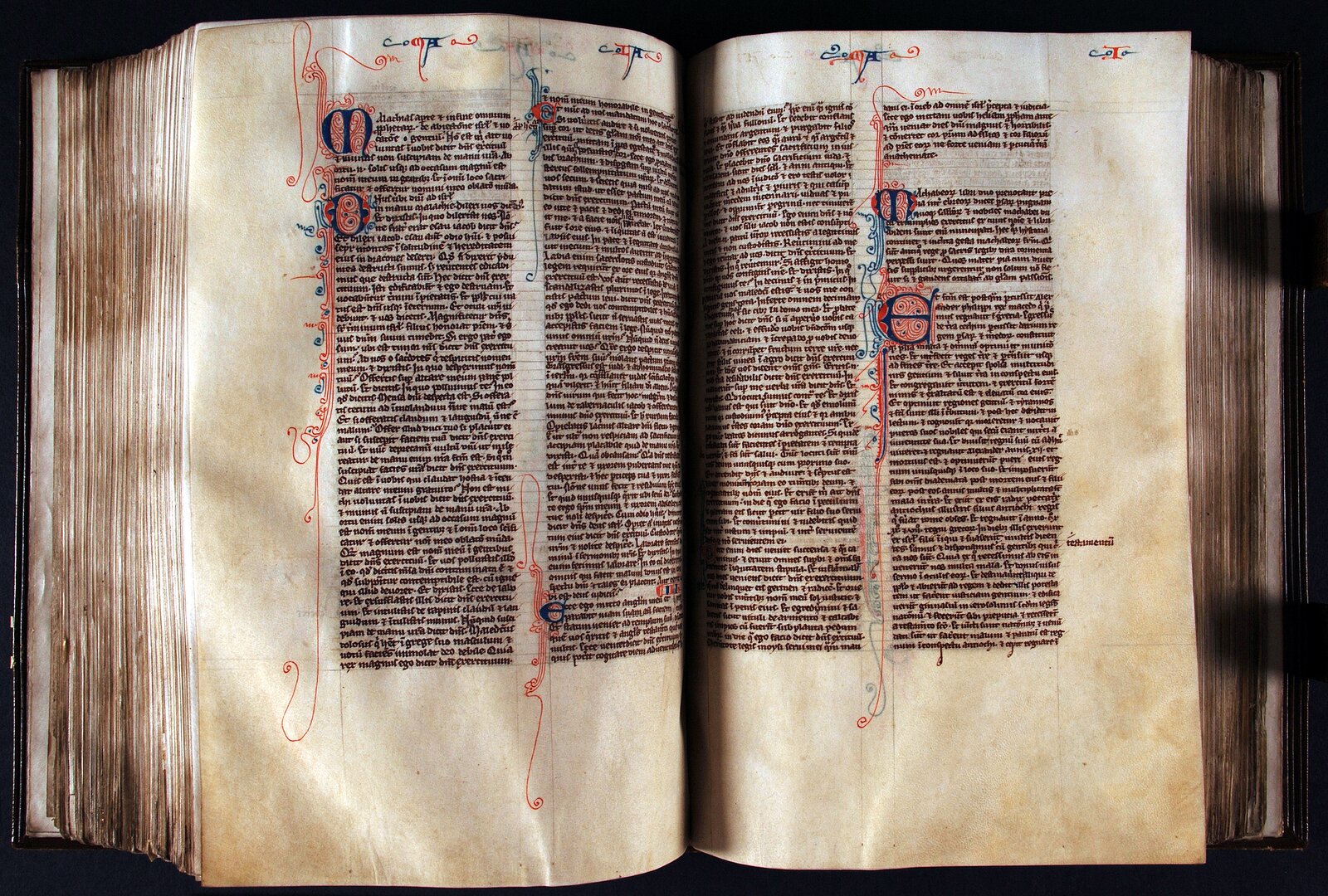Kolorowe zdjęcie przedstawia otwartą księgę. Jej strony są pożółkłe, nieco sfatygowane. Widoczne stronice są zapisane drobnym, równym, odręcznym pismem w dwóch kolumnach. Litera rozpoczynająca akapit jest większa i pięknie zdobiona ornamentami w kolorze niebieskim i czerwonym. Tło jest czarne.