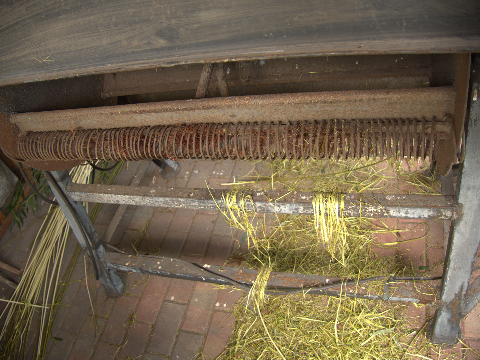 Zdjęcie przedstawia korowarkę mechaniczną. Urządzenie składa się z metalowej podstawy, bębnów korujących oraz drewnianej obudowy. Pod urządzeniem leżą fragmenty kory, a obok pręty okorowanej wikliny. 