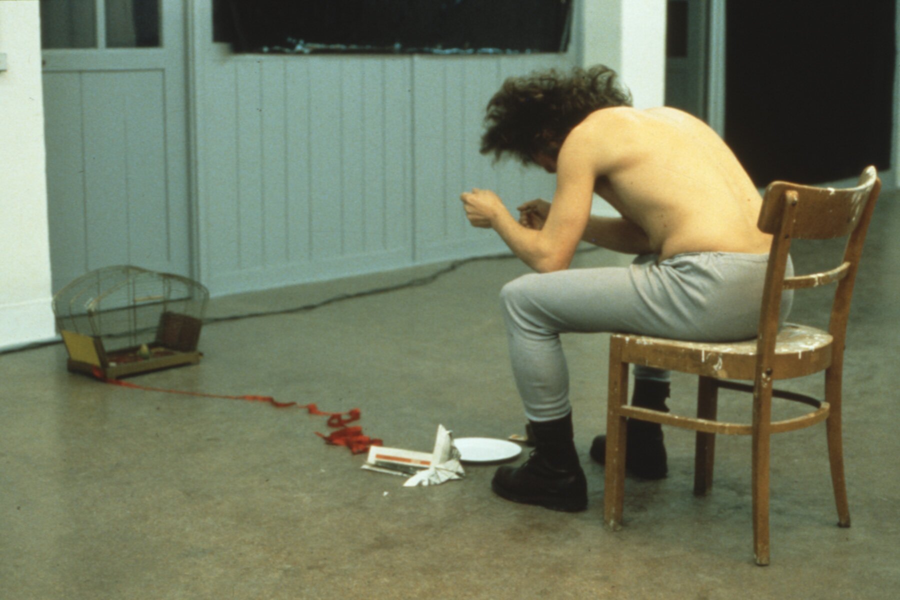 Zdjęcie przedstawia mężczyznę, siedzącego w pochylonej pozycji na krześle. Krzesło jest drewniane i pochlapane białą farbą. Mężczyzna ma zwichrzone włosy i ręce oparte łokciami na nogach. Ma na sobie szare spodnie dresowe i czarne, skórzane buty. Przed nim, na podłodze leży talerz i inne przedmioty. Dalej znajduje się czerwona wstążka, a następnie klatka z małym ptakiem. Performance odbywa sie w zamkniętym pomieszczeniu. Podłoga jest szara. Za klatką stoja szare drzwi.