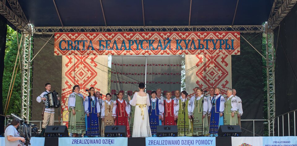 Zdjęcie przedstawia grupę kobiet i mężczyzn w kolorowych strojach ludowych, która stoi na scenie. Większość osób śpiewa do ustawionych przed nimi mikrofonów, mężczyzna po lewej stronie gra na akordeonie. Przed grupą stoi dyrygentka w białej sukni. Na scenie wisi wzorzysta tkanina z napisem w języku białoruskim.