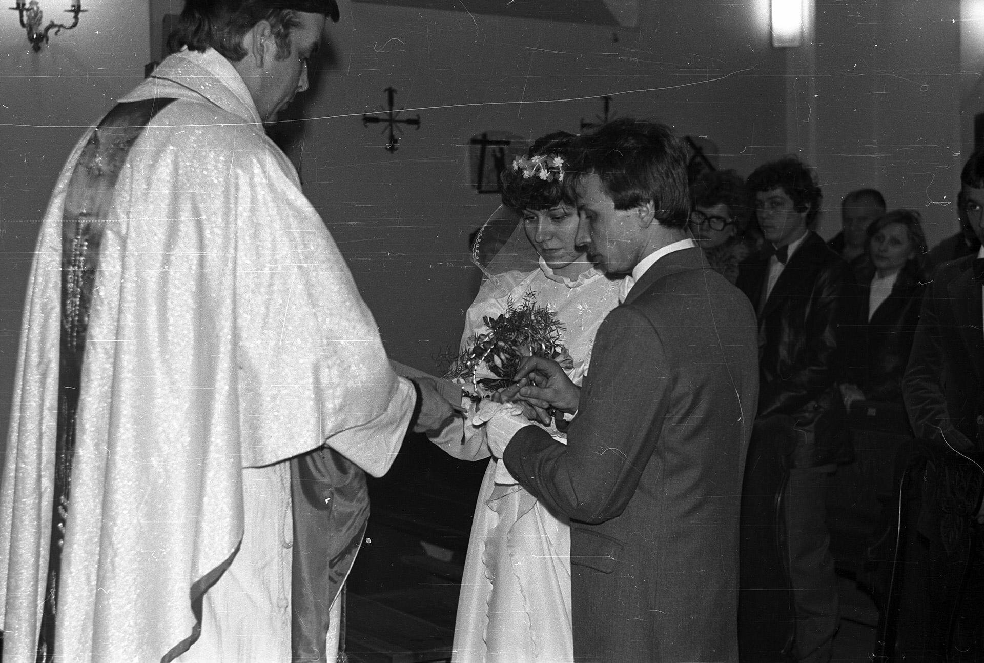 Zdjęcie przedstawia sakrament małżeństwa zawierany w kościele. Przed księdzem stoi młoda para. Młoda kobieta jest w białej sukni, na głowie ma wianek z przyczepionym do niego welonem. W dłoniach bukiet kwiatów. Mężczyzna jest w garniturze. Wkłada kobiecie obrączkę na serdeczny palec prawej dłoni. W tle stoją ludzie.