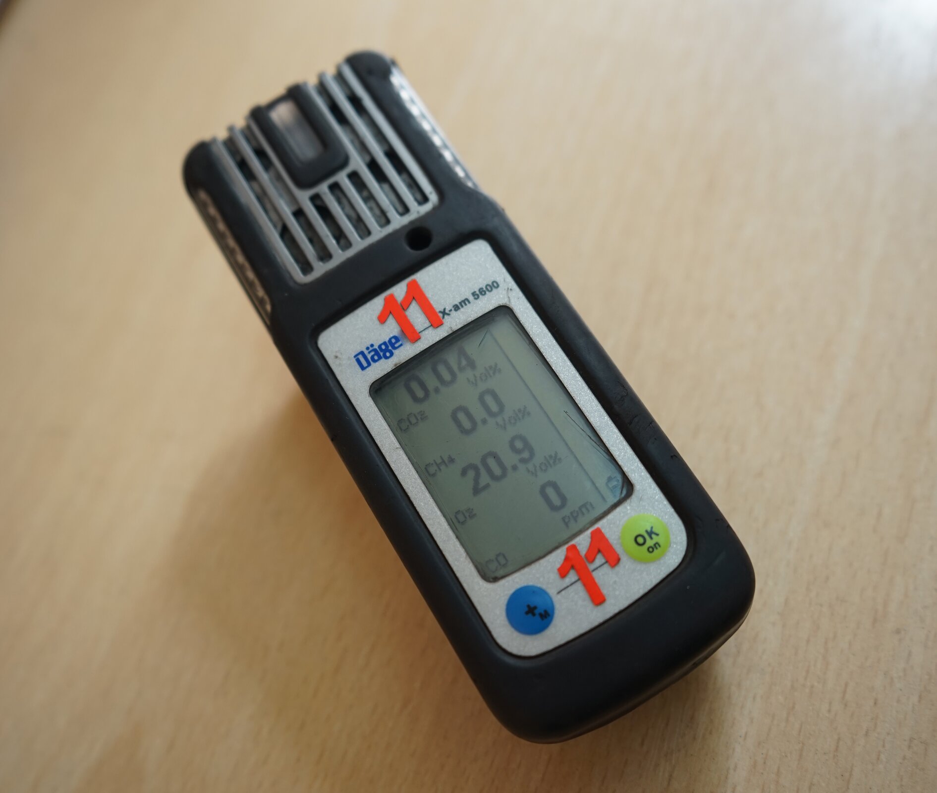 Zdjęcie przedstawia detektor uniwersalny, mogący wykryć od jednego do sześciu gazów. Detektor ma czarną obudowę o kształcie prostopadłościanu. Na przedniej ścianie obudowy znajduje się duży wyświetlacz, zajmujący ponad połowę powierzchni. Na ekranie wyświetlone są oznaczenia: dwutlenek węgla, metan, tlen, tlenek węgla. Przykładowe wyświetlane wartości: dwutlenek węgla – cztery setne, metan – zero, tlen - dwadzieścia i dziewięć dziesiątych, tlenek węgla – zero procent. Pod ekranem znajdują się dwa okrągłe przyciski. Po lewej stronie niebieski z symbolem plus. Po prawej stronie zielony z symbolem: ok. 