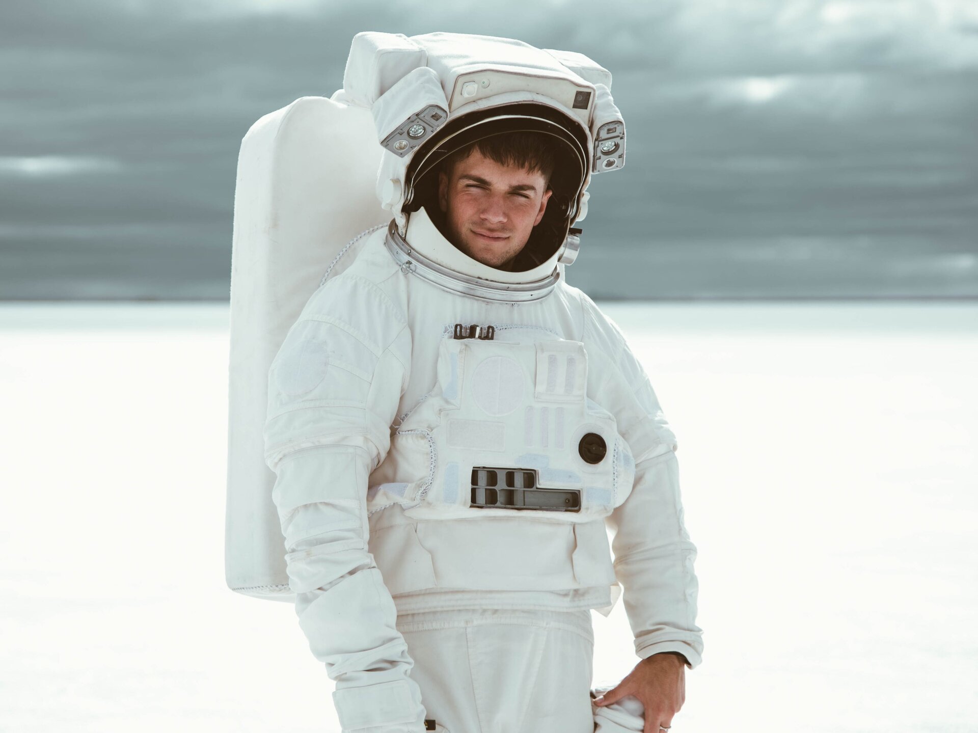 Zdjęcie przedstawia młodego mężczyznę ubranego w biały skafander i biały hełm z otwartą szybką na twarzy. Na plecach mężczyzna ma biały plecak kosmonauty z tlenem. W tle widoczna jest u dołu rozmyta biała powierzchnia, a u góry szara powierzchnia.
