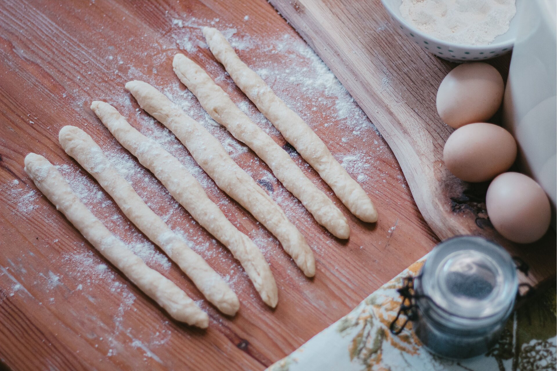 Zdjęcie przedstawia krok drugi i trzeci formowania chały kijowskiej. Ujęcie wykonane jest od góry pod skosem. W środkowej części grafiki widoczny jest drewniany ciemnobrązowy blat na którym leży sześć podłużnych i owalnych wałków jasnego ciasta. Oprószone są one mąką, która rozsypana jest również dookoła nich na blacie. W lewej górnej części zdjęcia znajduje się drewniana jasnobrązowa deska. W lewym górnym rogu, na desce stoi biała miska w niebieskie małe kropki. Obok miski po prawej stronie widoczne są trzy jajka za którymi dostrzec można biały wysoki kwadratowy pojemnik. W prawym dolnym rogu znajduje się biały obrus w zielone i żółte motywy roślinne. Na nim stoi szklany przezroczysty słoik z czarnym zatrzaskiem w środku wypełniony niebieskim makiem.