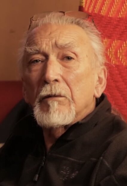 Kolorowe zdjęcie przedstawia portret mężczyzny w podeszłym wieku. Postać ma siwe, zaczesane do tyłu włosy, siwe wąsy, bródkę i krzaczaste, siwe brwi. Na głowę ma założone okulary. Ubrany jest w ciemną bluzę zapinaną na zamek błyskawiczny, pod szyją lekko rozpiętą. W tle znajduje się czerwono‑pomarańczowa, tkana w jodełkę zasłona. 