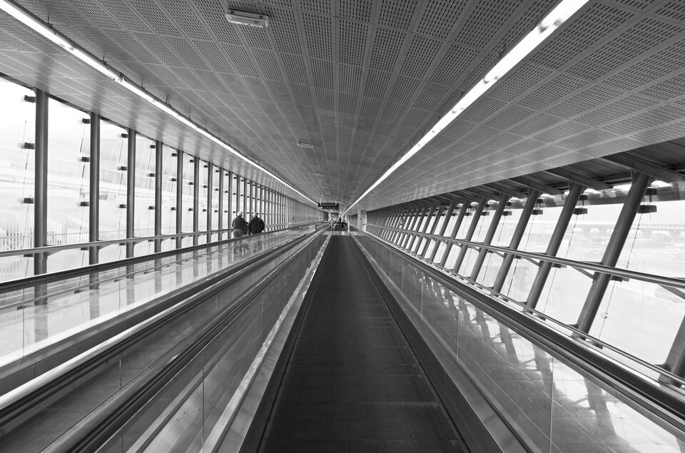 Fotografia Berta Kaufmanna przedstawia halę transferową na lotnisku. Zdjęcie w odcieniach szarości. Centralną część zajmuje ciemnoszary chodnik ze szklanymi barierkami po obu stronach. Po prawej stronie ściana ze szkła, przed nią metalowe wsporniki i pozioma barierka. Ściana jest lekko odchylona od pionu. Po lewej stronie również szklana ściana,prosta i wyższa, podzielona wspornikami. W oddali, po lewej stronie dwie postacie. Na suficie po prawej i lewej stronie ciąg świetlówek. Całość skupia się w jednym centralnym punkcie, w którym łączą się linie proste, poziome.