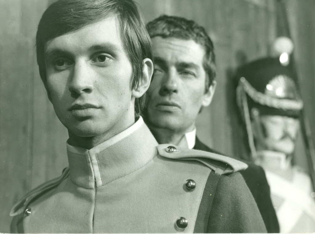 Zdjęcie przedstawia trzech mężczyzn. Na pierwszym planie znajduje się młodzieniec w mundurze wojskowym. Ma spokojną twarz. Za jego plecami stoi dojrzały mężczyzna w surducie. Na drugim planie znajduje się żołnierz w mundurze z wysoką czapką na głowie.