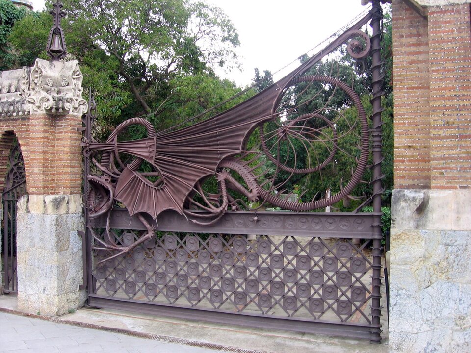 Ilustracja o kształcie poziomego prostokąta przedstawia wejście do pawilonów Guell. Brama wejściowa wykonana jest z kutego żelaza wraz i osadzona między słupami z kamienia i cegły. Dekorowana jest motywami fantastycznymi przypominającymi skrzydła smoka. Za brama widoczna jest część parku Guell bogata w roślinność. 