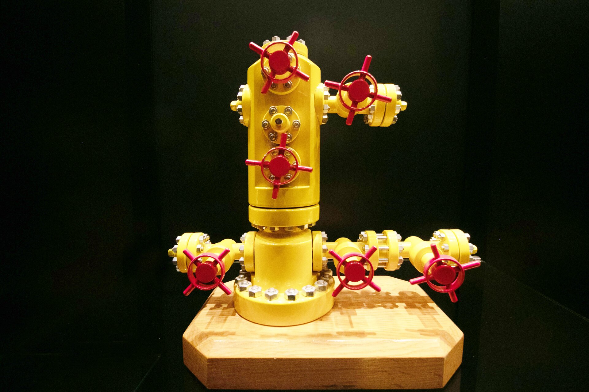 Na zdjęciu widać żółto‑czerwoną głowicę eksploatacyjną typu Solid Block, czyli urządzenia montowanego na szczycie odwiertu, w celu zamknięcia wylotu otworu wiertniczego, aby nie doprowadzić do niekontrolowanego ujścia węglowodorów.  Korpus głowicy składa się z dwóch elementów o kształcie cylindrycznym połączonych w połowie wysokości korpusu połączeniem kołnierzowym. Górny element korpusu jest dłuższy od dolnego. W dolnej części korpusu widoczne są po prawej i lewej stronie przyłącza rurowe. Przyłącze znajdujące się po prawej stronie składa się z odcinków rurowych na których osadzone są dwie zasuwy ślimakowe. Pokrętła zasuw są okrągłe w kolorze czerwonym. W środku pokrętła znajduje się dodatkowy czerwony okrągły element. Przez całe pokrętło przechodzą cienkie cylindryczne kształty tworzące znak: X. Kurek tworzy przez to kształt okręgu z opisanym na nim znakiem: X. Po drugiej stronie głowicy znajduje się przyłącze rurowe z jednym pokrętłem do zasuwy ślimakowej.W górnej części głowicy znajduje się jedno przyłącze rurowe. Przyłącze umieszczone jest po prawej stronie głowicy. Na pokrętle znajduje się zasuwa ślimakowa.W górnej części korpusu głowicy znajdują się dwa pokrętła do zasuwy ślimakowej. Pierwsze umieszczone jest poniżej krawędzi głównej głowicy a dolne powyżej połączenia kołnierzowego. Wszystkie pokrętła są takiego samego koloru, kształtu i wielkości jak w przypadku pokręteł do zasuw znajdujących się w dolnej części głowicy.Podstawą głowicy jest połączenie kołnierzowe.Głowica umieszczona jest na drewnianej sześciokątnej płycie. Tło za głowicą jest czarne.