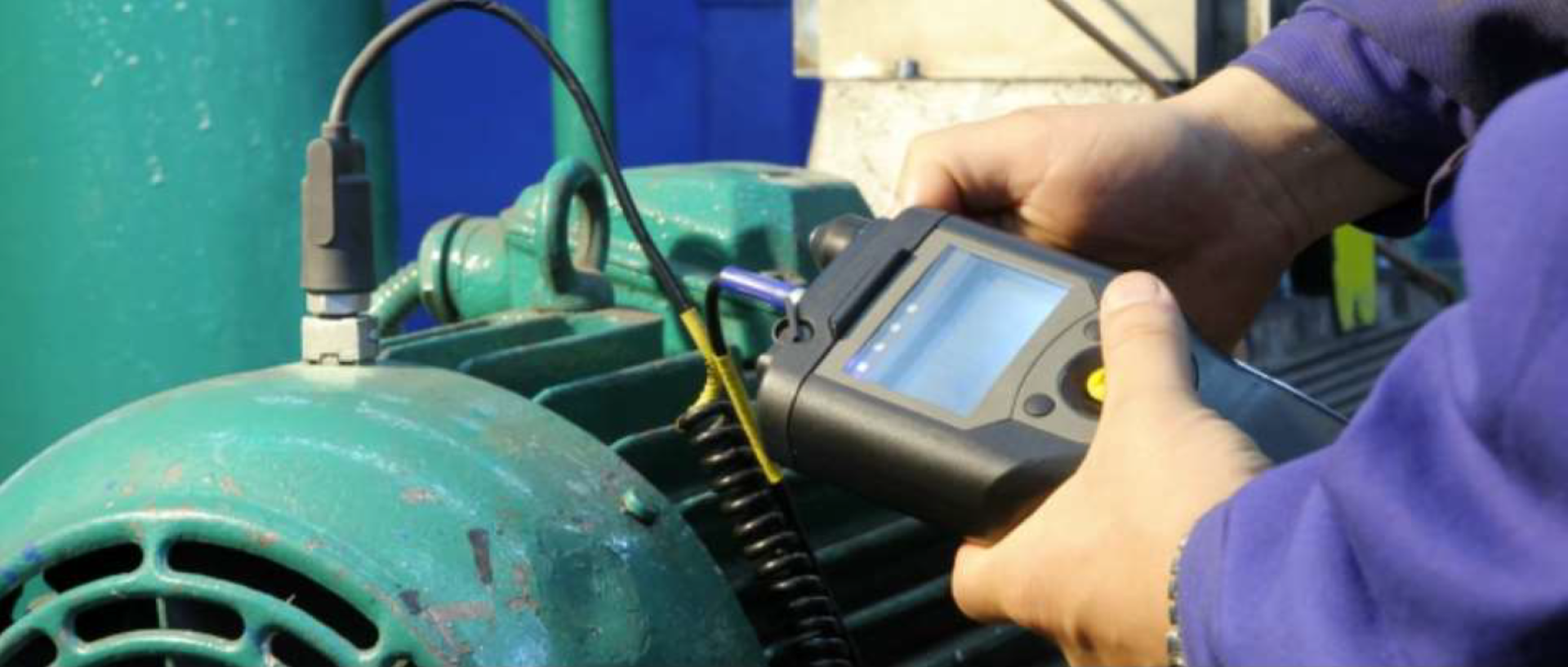 Na załączonym zdjęciu mężczyzna trzyma urządzenie, służące do pomiaru częstotliwości drgań silnika. Sprzęt jest podłączony do silnika.