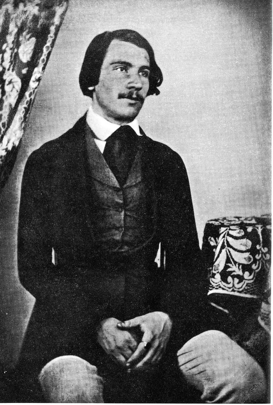 Fotografia przedstawia siedzącego, młodego mężczyznę, który patrzy w dal. Ma złożone między nogami dłonie, jest lekko uśmiechnięty. Jest ubrany we frak z kamizelką, pod którą znajduje się biała koszuka z zawiązanym krawatem. Mężczyzna ma czarne, proste włosy, które sięgają mu za ucho, jest gładko uczesany. Brwi grube, nos gruby szeroki, pod nim wąsik. W tle, z lewej strony znajduje się fragment wzorzystej zasłony. Z prawej strony stoi okrągły stolik z kolorowym obrusem.