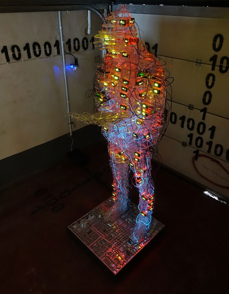 Prostokątna fotografia prezentuje rzeźbę przedstawiającą człowieka, który trzyma książkę. Posąg wykonany jest z wielu kabli oraz mechanizmów. Rzeźba podświetla się pomarańczowymi i żółtymi diodami. Ściany wokół posągu są białe z czarnymi kombinacjami cyfr 1 oraz 0.