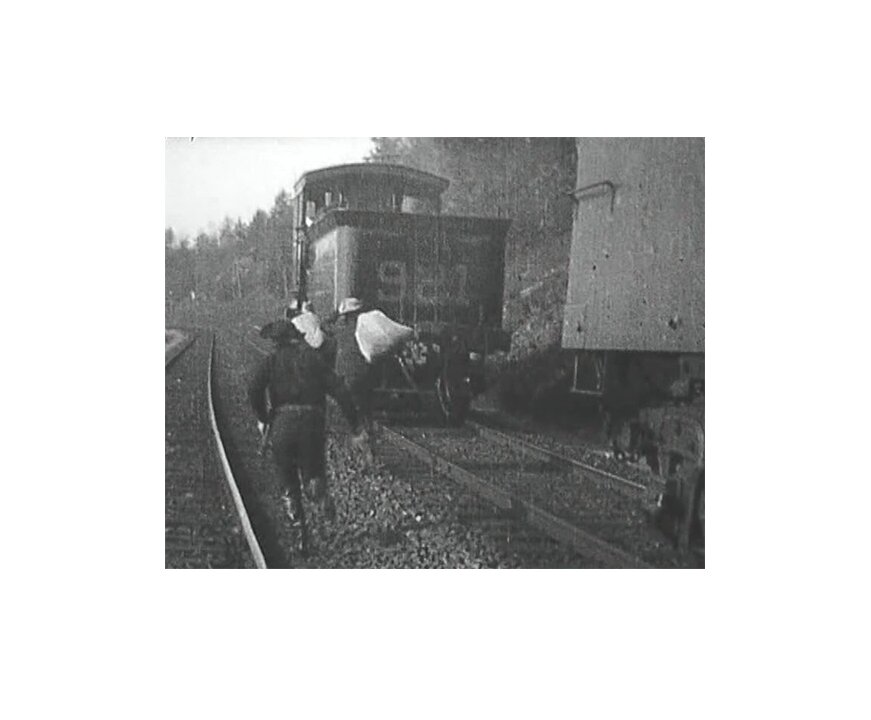 Kadr z czarno-białego filmu. Przedstawia trójkę mężczyzn w kowbojskich kapeluszach, którzy biegną wzdłuż jadącego lub stojącego na torze pociągu. Dwóch mężczyzn ma na plecach białe worki. Trzeci, biegnący za nimi, ma na sobie czarny strój i kapelusz. Z boku trzyma pistolet. Frame from black and white film. It presents three men in cowboy hats who are running along a train or standing on a track. Two men have white sacks on their backs. The third one, running after them, is wearing a black outfit and a hat. Gun is on the side.