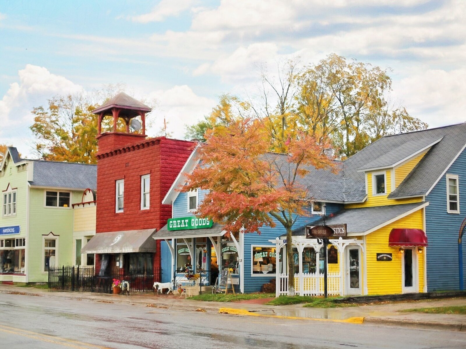Zdjęcie przedstawia niewysokie, kolorowe budynki stojące przy ulicy. Na frontach budynków widoczne są witryny sklepowe i markizy, a nad wejściami napisy w języku angielskim, na przykład: GREAT GOODS.