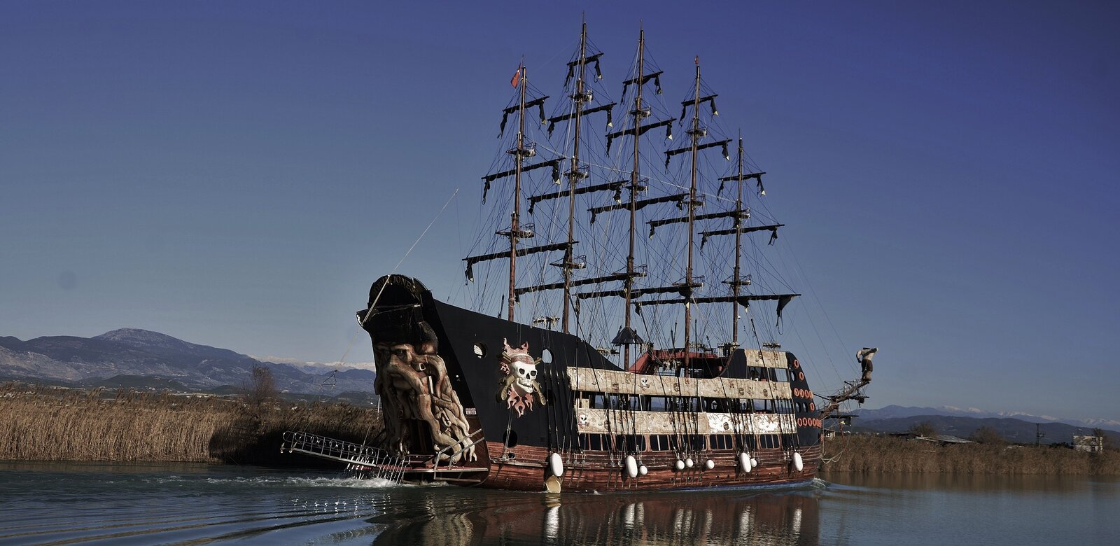 Na zdjęciu znajduje się drewniany statek o dwóch zabudowanych pokładach i trzecim odkrytym, który wolno płynie po spokojnej tafli wody. Od szczytu znajduje się duża, przestrzenna ozdoba w postaci głowy mężczyzny – postać ma macki ośmiornicy, które układają się w kształt wąsów, brody i włosów, oraz srogie spojrzenie. Na głowie znajduje się czapka piracka. Nad postacią jest nazwa statku Davy Jones Na burcie, po lewej stronie, znajduje się dekoracja w postaci dużej trupiej czaszki z bandaną na czole, zza której wystają dwa skrzyżowane pistolety i płomienie. Wzdłuż burty ciągną się dwie szerokie listwy, na których namalowano mapy, trupie czaszki, skrzyżowane piszczele, szable. Statek ma pięć masztów, żagle są zwinięte. Na pierwszym od lewej maszcie powiewa mała chorągiewka. W tle widać niskie nabrzeże porośnięte zrudziałą trawą, w oddali widnieją szczyty gór. Niebo jest bezchmurne.