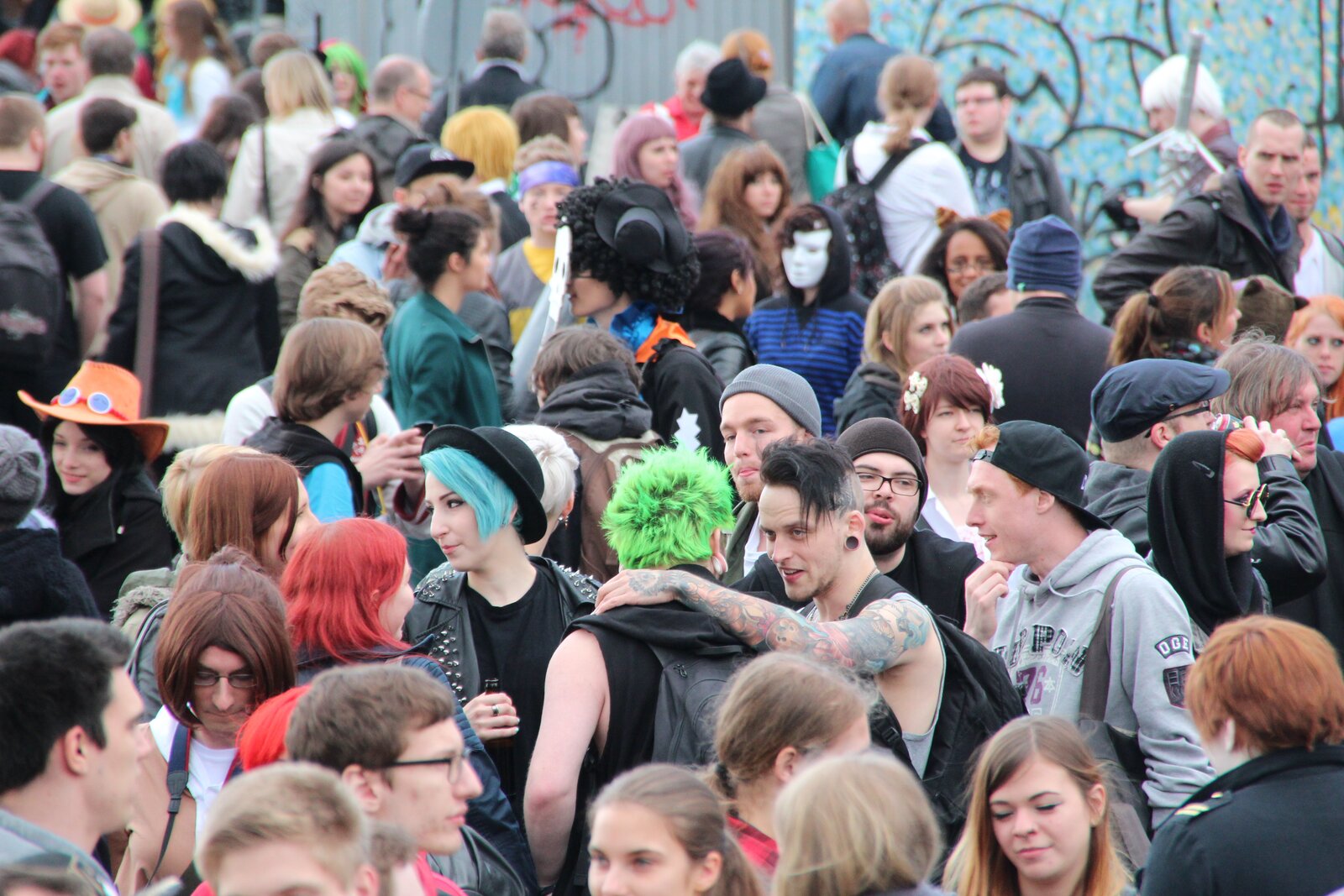 Zdjęcie przedstawia tłum ludzi. Część z nich m a włosy nienaturalnego koloru, inni mają maski, kolorowe kapelusze.