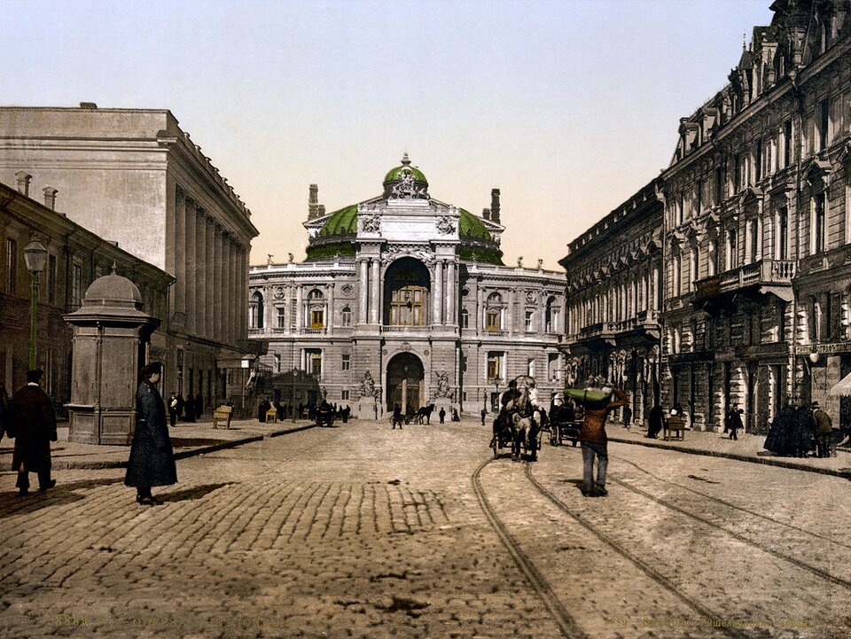 Grafika przedstawia widok na dwupiętrowy budynek, którego szczyt wieńczy zielona kopuła. Do budynku prowadzi pokryta kostką brukową droga, po obu jej stronach widać kamienice. Na ulicy znajdują się ludzie, na drugim planie są również powozy konne.