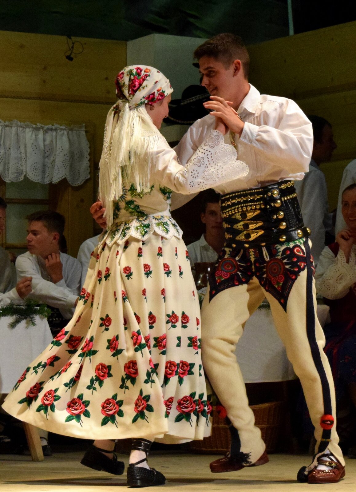 Fotografia przedstawia prezentacje tradycyjnego stroju podhalańskiego. Na fotografii znajduje się tańcząca para. Kobieta ubrana jest w czarne buty, długą białą suknię ozdobioną kwiatowym wzorem, białą haftowaną koszulę oraz kwiatową chustę na głowie. Mężczyzna ubrany jest w brązowe buty, jasne spodnie ze zdobieniami oraz białą koszulę. W tle obrazka znajdują się mężczyźni ubrani w białe koszule.