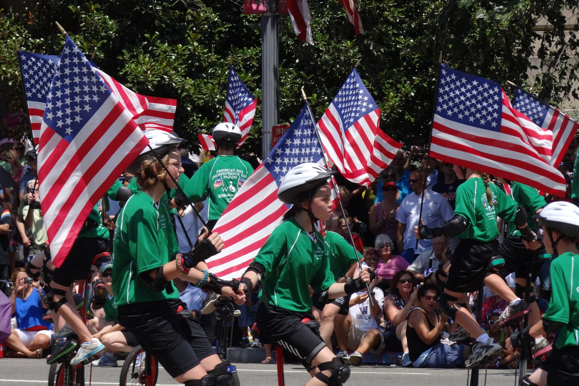 Zdjęcie przedstawia na pierwszym planie grupę młodych osób, ubranych w takie same zielone koszulki, ochraniacze na kolana i ręce, białe kaski, trzymających amerykańskie flagi w rękach. Na koszulkach widnieje napis w języku angielskim: America's 2012 Independence Day Parade. Poniżej widoczne jest jakieś logo złożone m.in. z dwóch flag. Na pierwszym planie zdjęcia, pośrodku, widoczni są chłopak i dziewczyna jadący na rolkach w prawą stronę. W tle widoczne są postacie wykonujące akrobacje na rowerach. Za nimi widoczny jest tłum stojących i siedzących na ziemi widzów.