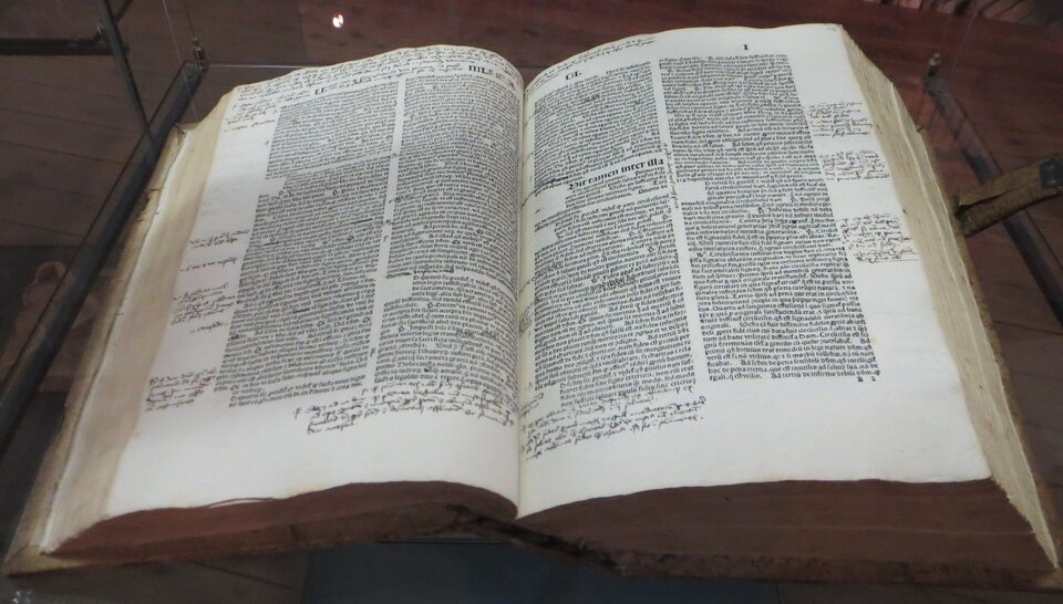 Zdjęcie przedstawia otwartą księgę w szklanej gablocie. Tekst na stronach zapisany jest w dwóch kolumnach. Ma marginesach liczne, odręczne notatki.