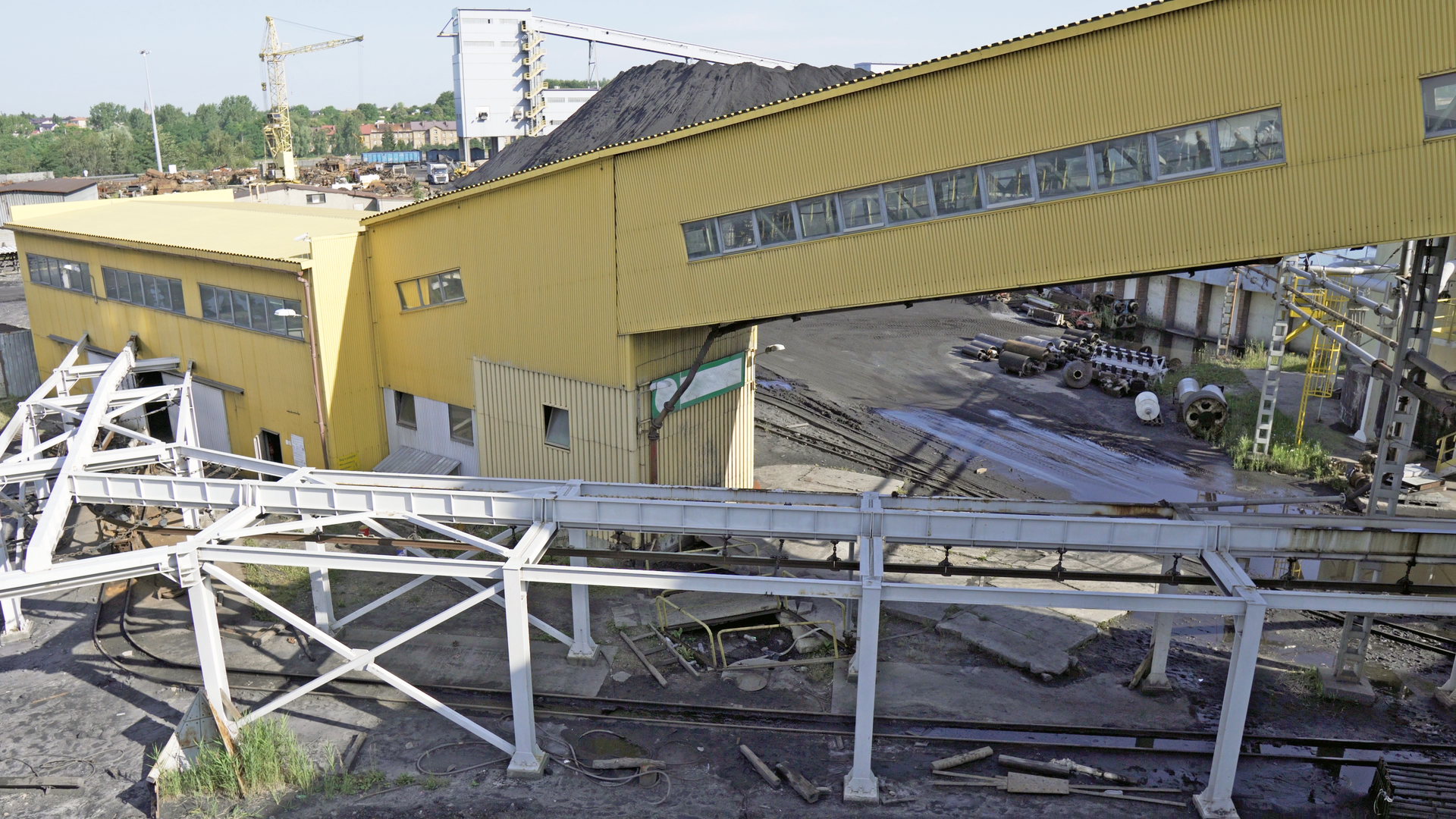 Na zdjęciu widoczny jest teren przemysłowy pokazany z lotu ptaka. Centralnym elementem zdjęcia jest żółty łącznik. Łącznik jest o kształcie prostopadłościanu znajdującego się pod kątem. W łączniku widoczne są okna. Przez okna widać prawdopodobnie przenośnik taśmowy.Na terenie placu widać części urządzeń oraz metalową konstrukcję przypominającą ramę przenośnika.