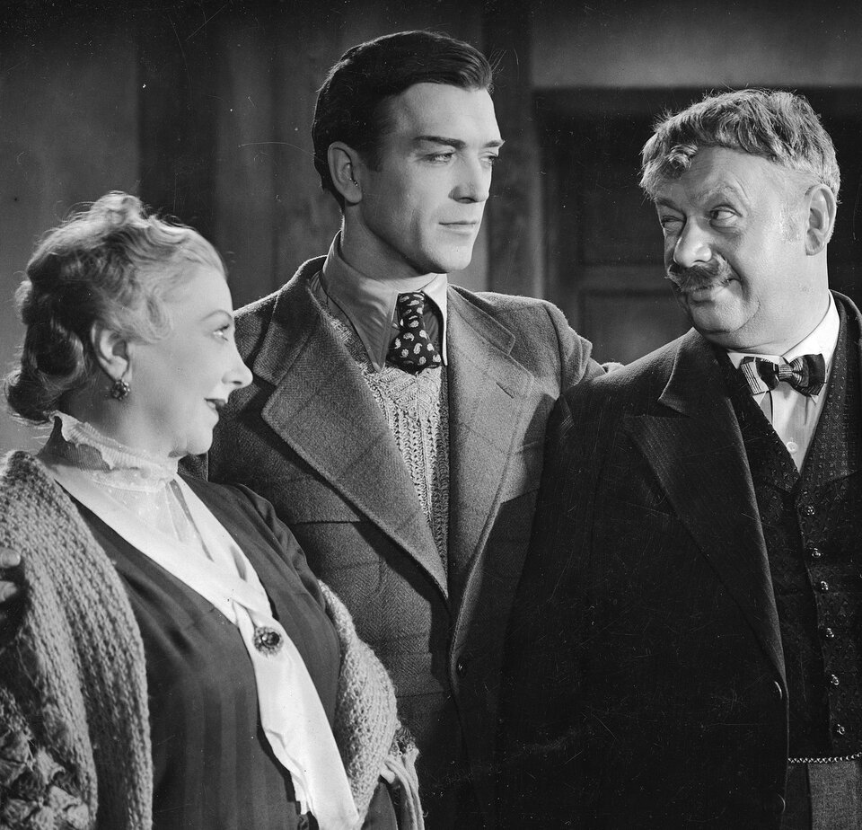 Fotografia przedstawiająca troje ludzi w różnym wieku. Po lewej stronie stoi dojrzała kobieta. Ukazuje prawy profil. Zaprezentowana jest od pasa w górę. Kobieta ma owalną twarz, cienkie brwi, zwężone oczy, zadarty nos i otwarte, wąskie usta. Ma zaczesane do tyłu, falujące włosy. Uszy kobiety zdobią małe kolczyki z kamieniem. Kobieta ubrana jest w bluzkę z kołnierzykiem wykończonym falbanką i w żakiet. Przez ramiona ma zawieszony szal wykonany na drutach. Do bluzki ma przywieszoną broszkę. Pośrodku stoi młody, wysoki mężczyzna. Postać ukazuje prawy półprofil. Ma owalną twarz. Jego włosy są krótkie, zaczesane do tyłu. Ma gładkie czoło, ciemne, wyraziste brwi. Jego nos jest prosty. Ma wąskie usta. Patrzy w prawą stronę. Mężczyzna jest ubrany w koszulę, kamizelkę robioną na drutach i marynarkę z wzorem w dużą kratę. Na szyi ma zawiązany krawat. Po prawej stronie stoi dojrzały mężczyzna. Ma owalną twarz. Jego włosy są krótkie, obcięte równo w kształt miski. Na czoło mężczyzny opada kręcony pukiel włosów. Ma jasne, uniesione brwi. Jego nos jest prosty. Ma sumiaste wąsy i zaokrągloną brodę. Patrzy w lewą stronę. Mężczyzna jest ubrany w koszulę, kamizelkę i marynarkę. Na szyi ma zawiązaną muchę. W tle znajduje się ściana i drzwi.