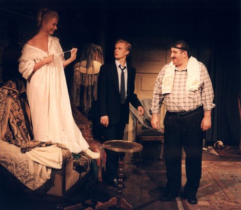 Zdjęcie ze spektaklu pt. "Tango". W pokoju, który sprawia wrażenie zagraconego, znajdują się: po lewej stronie kanapa z porozrzucanymi ubraniami, w lewym rogu stojąca lampa przykryta chustą, obok krzesło, w tle drzwi. Na środku mały okrągły stolik. Wszystko w brązowo‑beżowych barwach. Na łóżku stoi roześmiana kobieta, która ubrana jest w długą  białą  koszulę nocną. Młody mężczyzna w czarnym garniturze patrzy na nią z zaskoczeniem. Drugi mężczyzna, otyły, z wąsikiem, w koszuli w kratkę i ścierką położną na szyi, jest rozbawiony. 