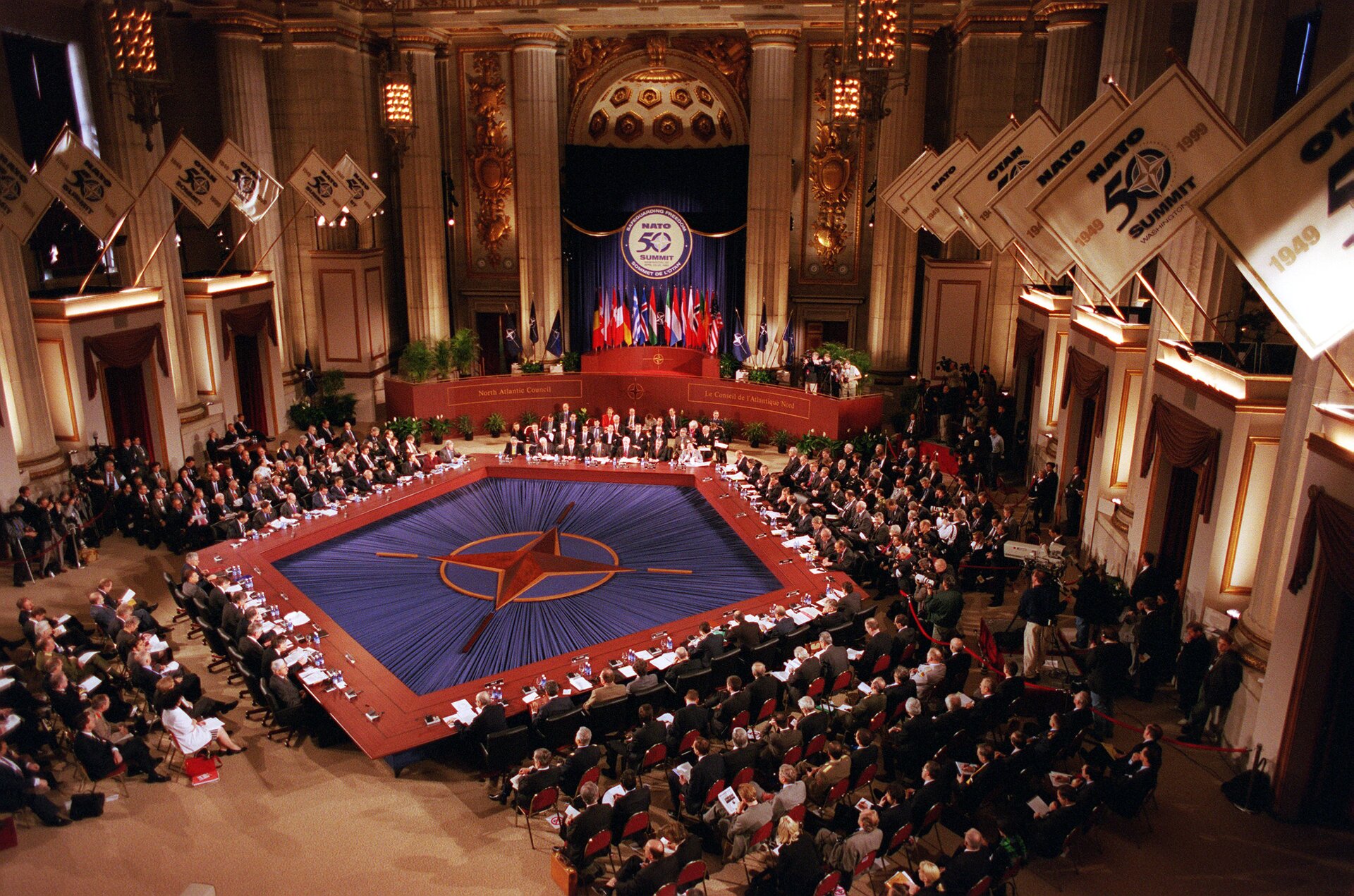 Zdjęcie przedstawia szczyt NATO, zorganizowany w Waszyngtonie w USA w dn. 23‑25 kwietnia 1999.  Szczyt został zwołany m.in. w celu uczczenia 50 rocznicy powołania Paktu Północnoatlantyckiego - w części centralnej widoczny napis z cyfrą 50 i flagi państw, które przynależą do NATO. Poniżej  podest, na którym widoczni są ludzie i poustawiane doniczki z krzewami. W centralnej części widoczna flaga NATO (w odcieniu błękitu, symbolizuje Ocean Atlantycki; pierścień symbolizujący jedność oraz na środku różę kierunkową oznaczającą bezpieczeństwo). Jest rozłożona, tworzy pięciokąt, wokół którego siedzą zaproszeni przedstawiciele państw NATO. Pozostali goście siedzą po bokach sali. Po bokach sali znajduję się filary pomiędzy którymi na wysokości pierwszego piętra są balkony. Na nich umiejscowione są flagi, które zwisają w kierunku środka sali. Na flagach znajdują się daty 1949,1999, oraz napisy NATO, Summit.