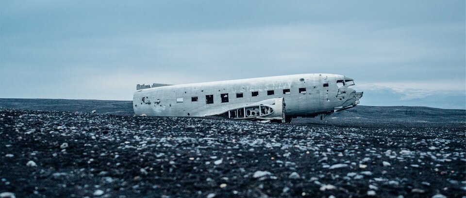 Zdjęcie przedstawia leżący na pustkowiu wrak samolotu. Maszyna ma tylko dziób, część przednią samolotu i dziurawy silnik z boku. Na ścianie samolotu widoczne jest osiem otworów po oknach i dwa otwory na dziobie, które były oknami dla pilotów. Leży na czarnej ziemi, na której widoczne są małe, białe fragmenty samolotu. Zdjęcie zostało zrobione w pochmurny dzień – kolorystyka zdjęcia jest utrzymana w szarościach.