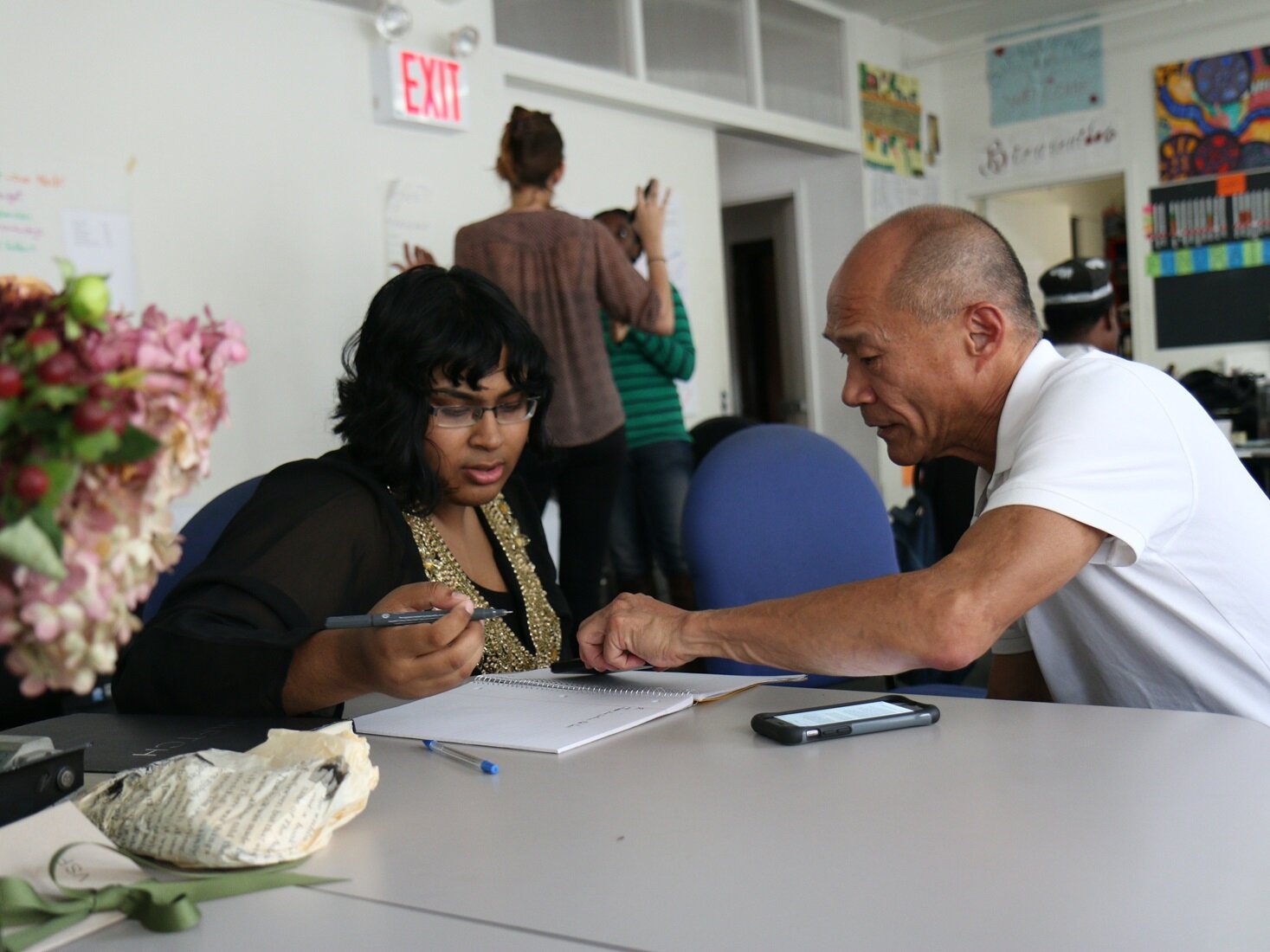 Zdjęcie przedstawia dwie osoby siedzące prze stole, na którym leży otwarty zeszyt, a obok długopisy i telefon. Kobieta w średnim wieku siedzi po lewej i ma skierowany wzrok na zeszyt, a w prawej ręce trzyma długopis. Starszy mężczyzna siedzący obok niej palcem wskazującym lewej ręki pokazuje coś w zeszycie. 