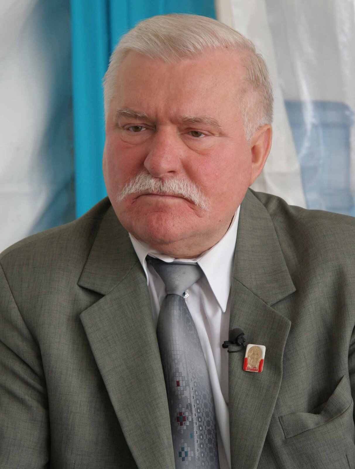 Na zdjęciu znajduje się prezydent Lech Wałęsa. Starszy mężczyzna z siwymi włosami oraz wąsami. Jest ubrany w garnitur. W klapie marynarki wpięty ma znaczek z wizerunkiem Matki Boskiej.