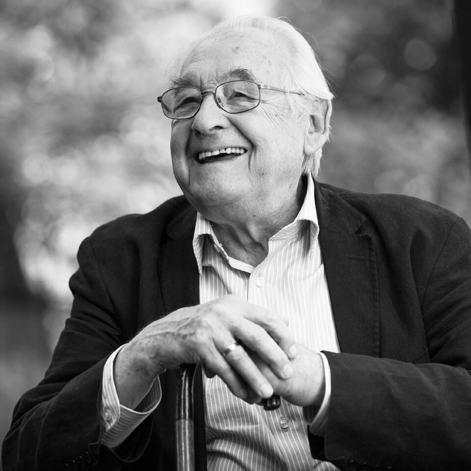 Fotografia przedstawia: Uśmiechnięty reżyser Andrzej Wajda. Siedzi oparty o laskę. Fotografia jest czarno‑biała. Reżyser ma pogodną twarz.