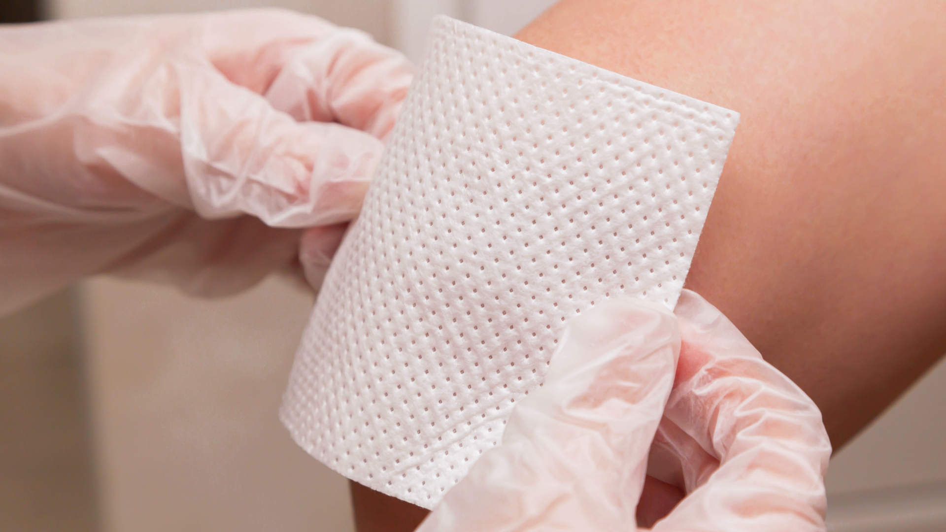 Zdjęcie przedstawia dłonie w gumowych rękawiczkach przykładające innej osobie opatrunek. 