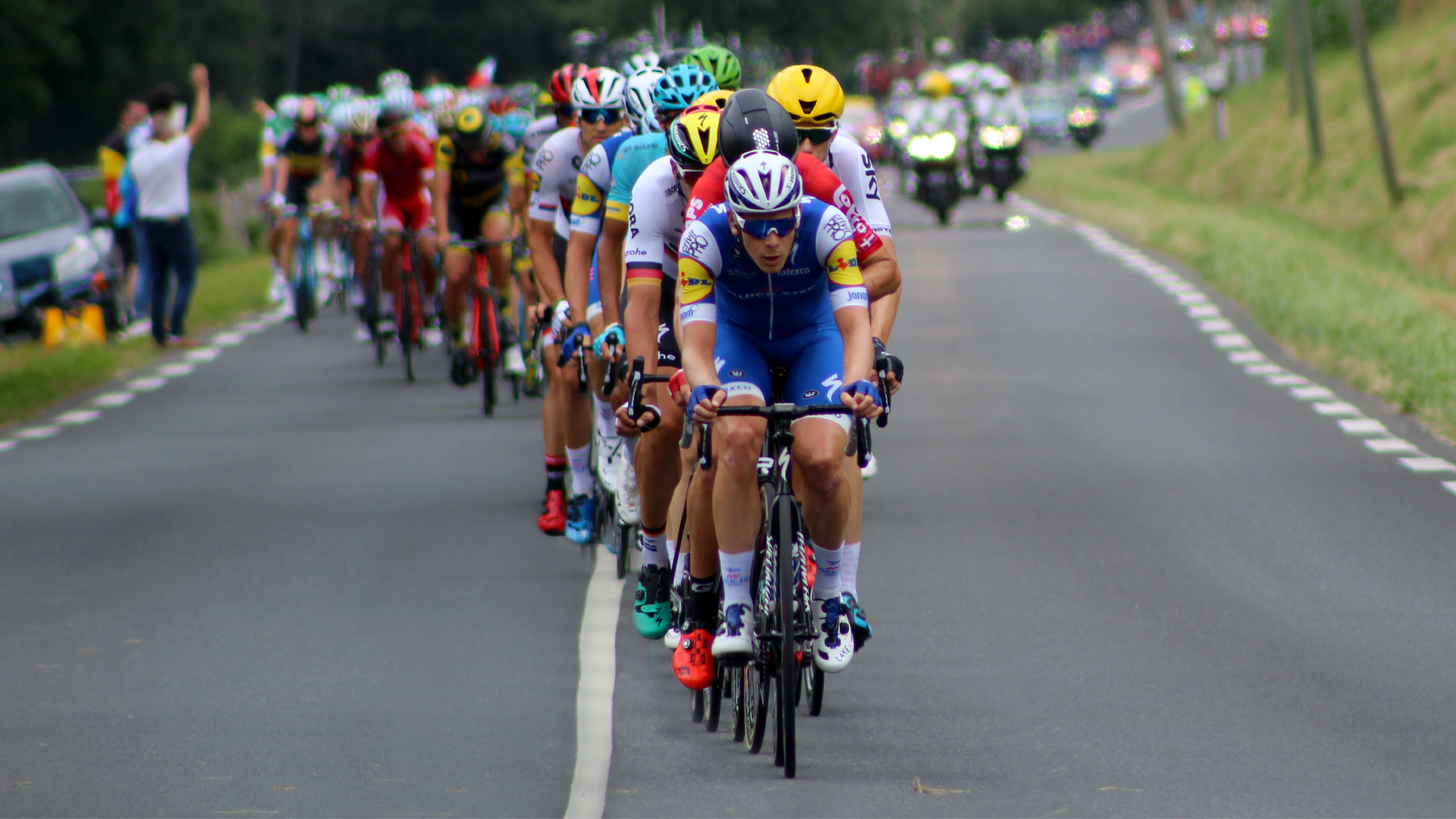Zdjęcie przedstawia peleton kolarzy, jadących drogą. W tle widoczne są motocykle z obsługi wyścigu. Poza drogą widocznych jest kilka osób oraz zaparkowany na poboczu samochód.