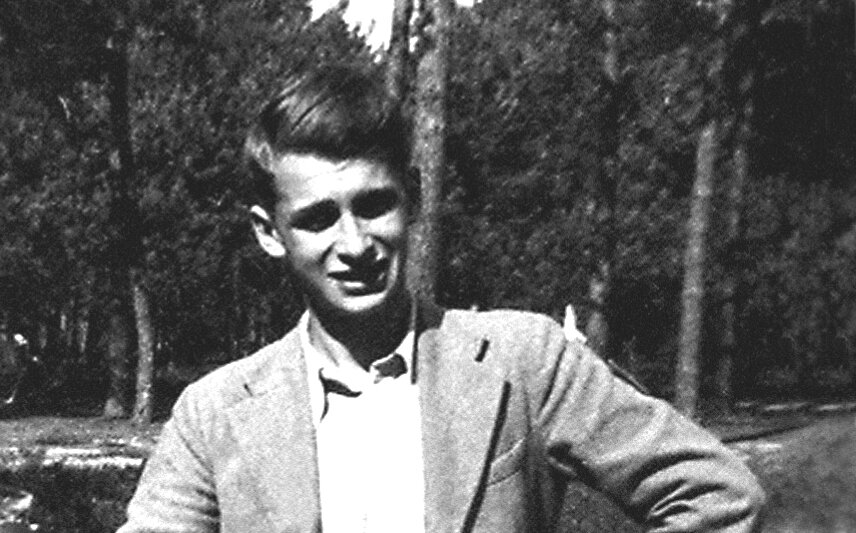 Ilustracja przedstawia czarno‑białe zdjęcie młodego mężczyzny w marynarce i białej koszuli. Mężczyzna jest szczupły, ma pociągłą twarz i bujne, ciemne włosy. W tle, za jego plecami, znajdują się drzewa.