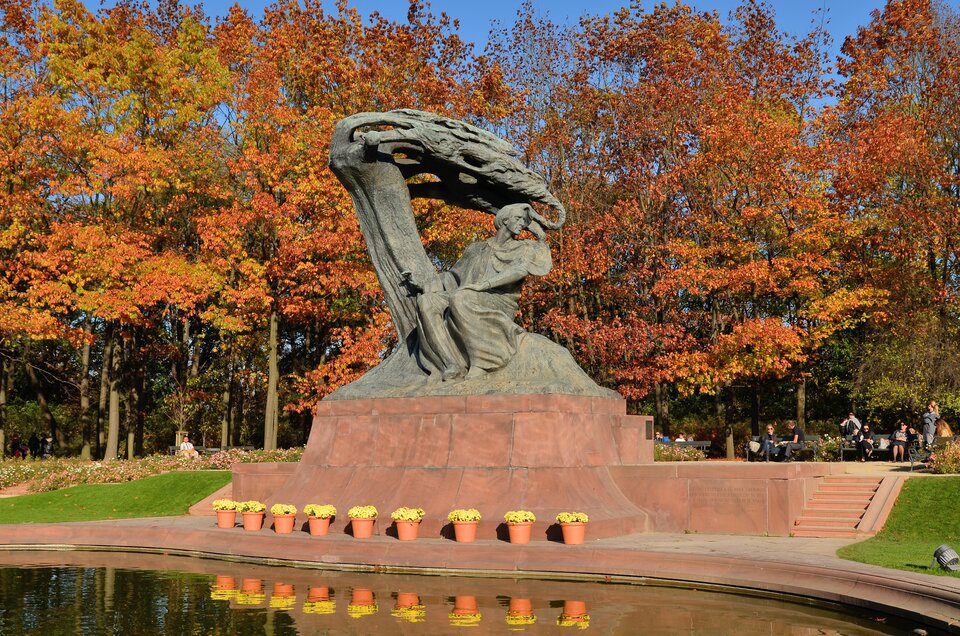 Zdjęcie przedstawia pomnik Chopina w stylu secesyjnym, znajdujący się w Łazienkach Królewskich w Warszawie – projekt Wacława Szymanowskiego. Pod pomnikiem stoją żółte kwiaty. Z tyłu, za pomnikiem widać jesienny żółto‑pomarańczowy krajobraz drzew. Z przodu akwen wodny.