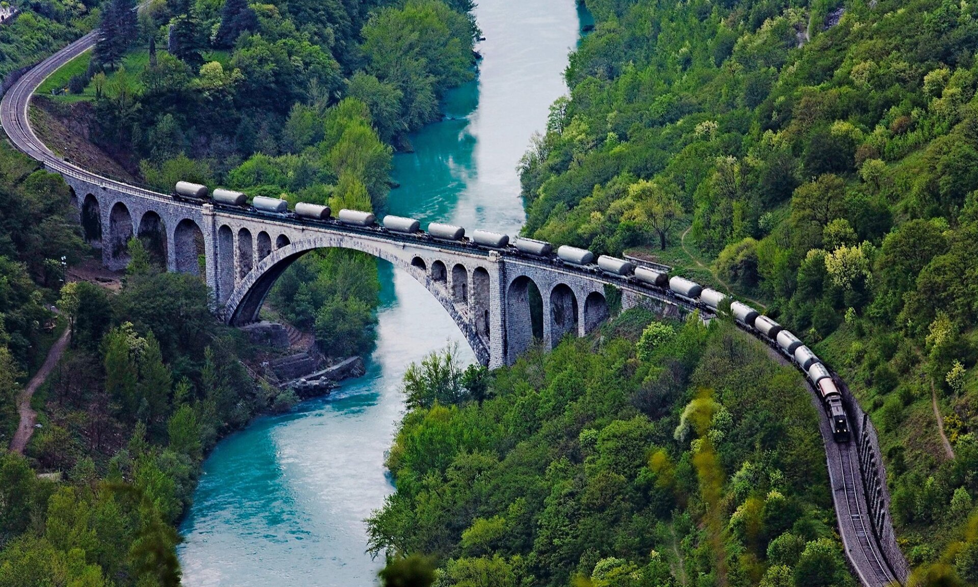 Zdjęcie ukazuje most łukowy widziany z perspektywy lotu ptaka. Na moście przemieszcza się pociąg z wagonami cysternowymi. Pod konstrukcją mostu rozpościera się błękitna rzeka, a wokół niej rosną drzewa. Most oparty jest na jednym głównym łuku pośrodku oraz kilku mniejszych łukach po bokach.