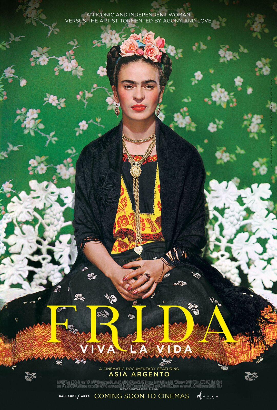 Ilustracja przedstawia plakat promujący film pod tytułem: „Frida: Viva la Vida” w reżyserii Giovanniego Troilo. Plakat przedstawia kobietę w średnim wieku o ciemnych włosach spiętych w kok, który został przyozdobiony różowymi kwiatami. Kobieta spogląda się prosto w oczy widza, ma pomalowane na pomarańczowo policzki oraz monobrew. Ubrana jest w długą czarną suknię z detalem w postaci białych kwiatków oraz pomarańczowego pasa biegnącego na wysokości łydek. Górna część sukni posiada kontrastowe, żółto‑pomarańczowe wzory kwiatowe. Kobieta ma duże, wiszące, złote kolczyki, duży pierścionek z kamieniem oraz masywny, złoty łańcuszek, sięgający do pasa. Kobieta siedzi na tle zielonej płachty przyozdobionej białymi kwiatami.