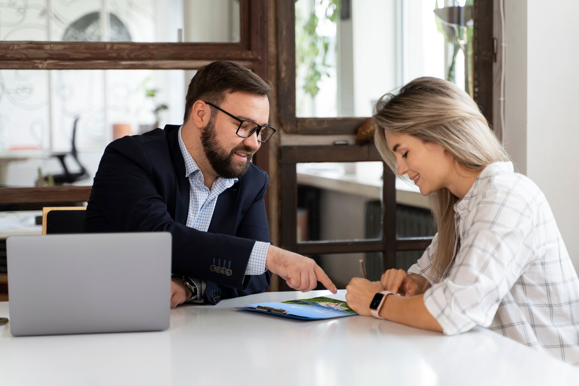 Na zdjęciu widać dwoje ludzi. Kobieta ubrana jest w białą koszulę w szarą kratę, ma blond włosy, jest lekko uśmiechnięta. Pisze coś na kolorowym zdjęciu. Mężczyzna ubrany jest w granatową marynarkę i koszulę w biało‑niebieską kratę, na twarzy ma delikatny zarost, grube, czarne okulary i ciemne włosy. Pokazuje kobiecie palcem coś na zdjęciu. Przed nim na stole stoi otwarty, srebrny laptop. W tle przeszklona ściana z drewnianymi ramkami, za nią kolejne pomieszczenie. 