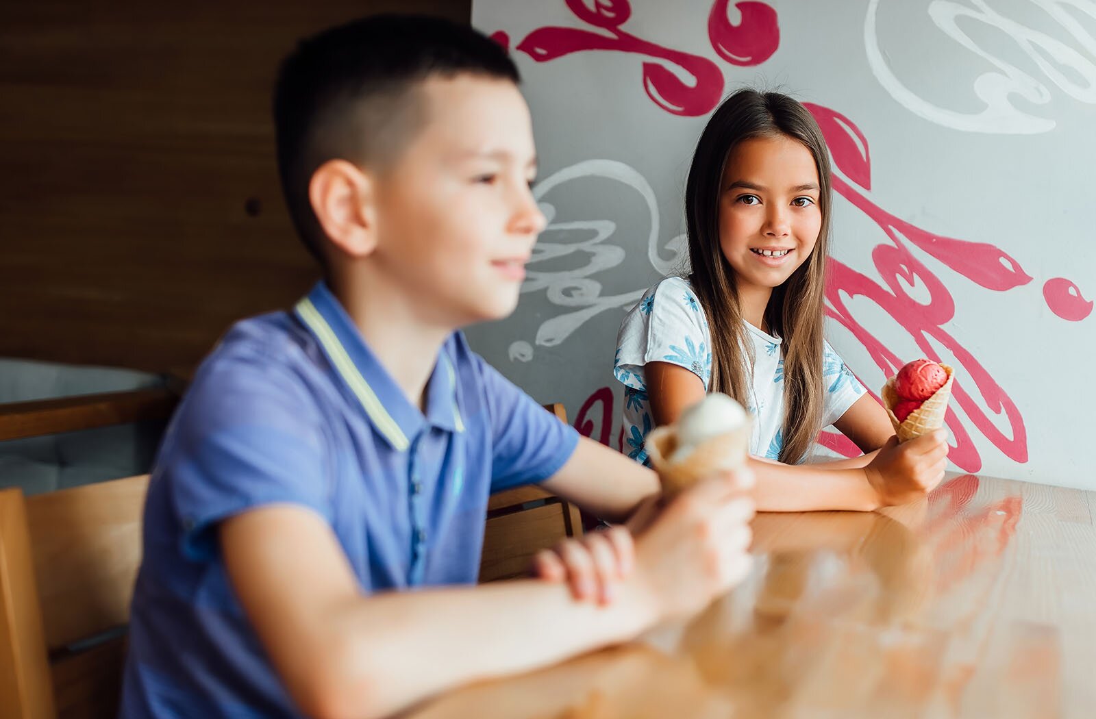 Zdjęcie przedstawia dwoje dzieci w wieku szkolnym siedzących przy stoliku. Oboje trzymają w dłoni lody.