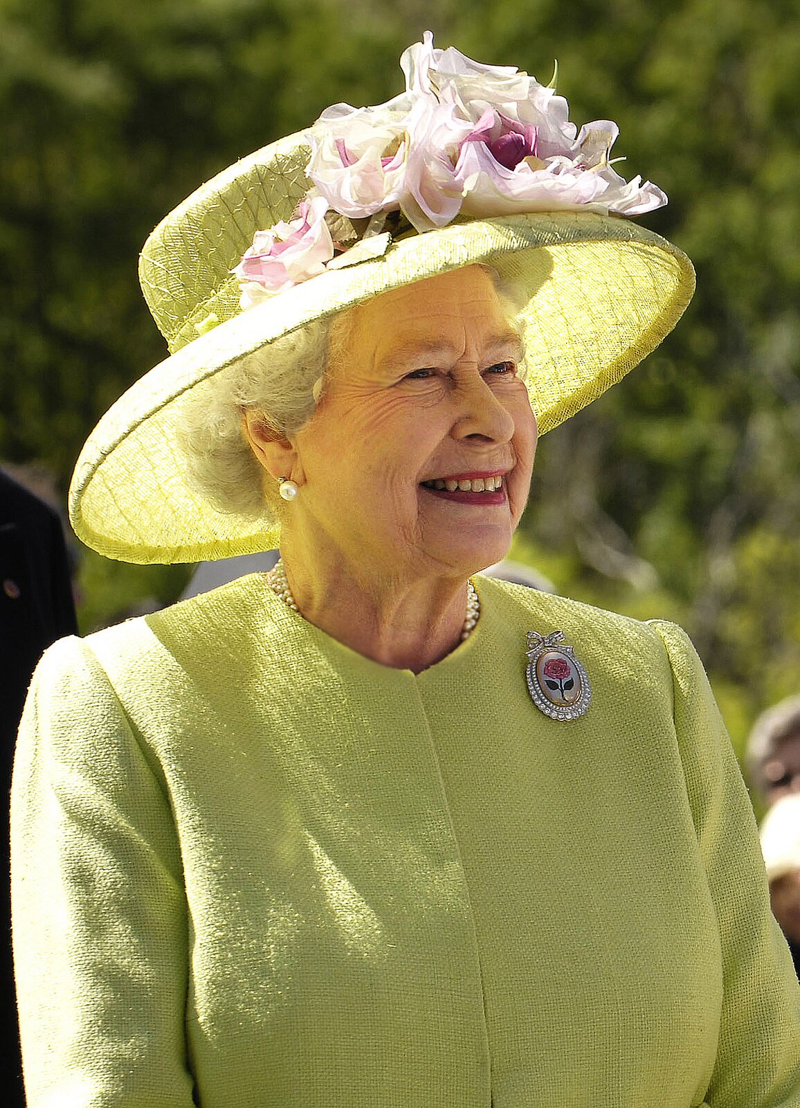 Zdjęcie przedstawia starszą, siwowłosą kobietę. To królowa Wielkiej Brytanii Elżbieta II. Jest ubrana w garsonkę, a na głowie ma kapelusz z dużym rondem, do którego dopięte są kwiaty.
