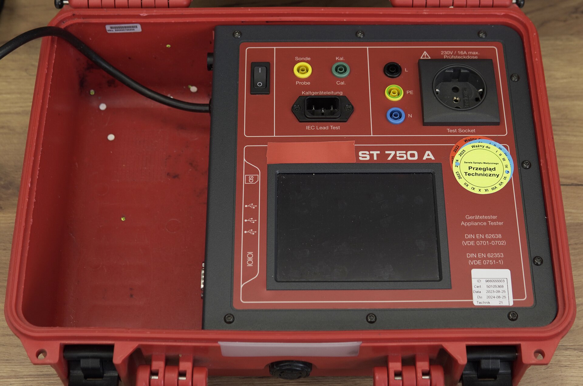 Zdjęcie przedstawia miernik bezpieczeństwa elektrycznego w perspektywie od góry. Urządzenie znajduje się w plastikowej, czerwonej skrzyni. Na górnej ścianie obudowy znajduje się włącznik, ekran oraz gniazda do podłączenia innych urządzeń. 