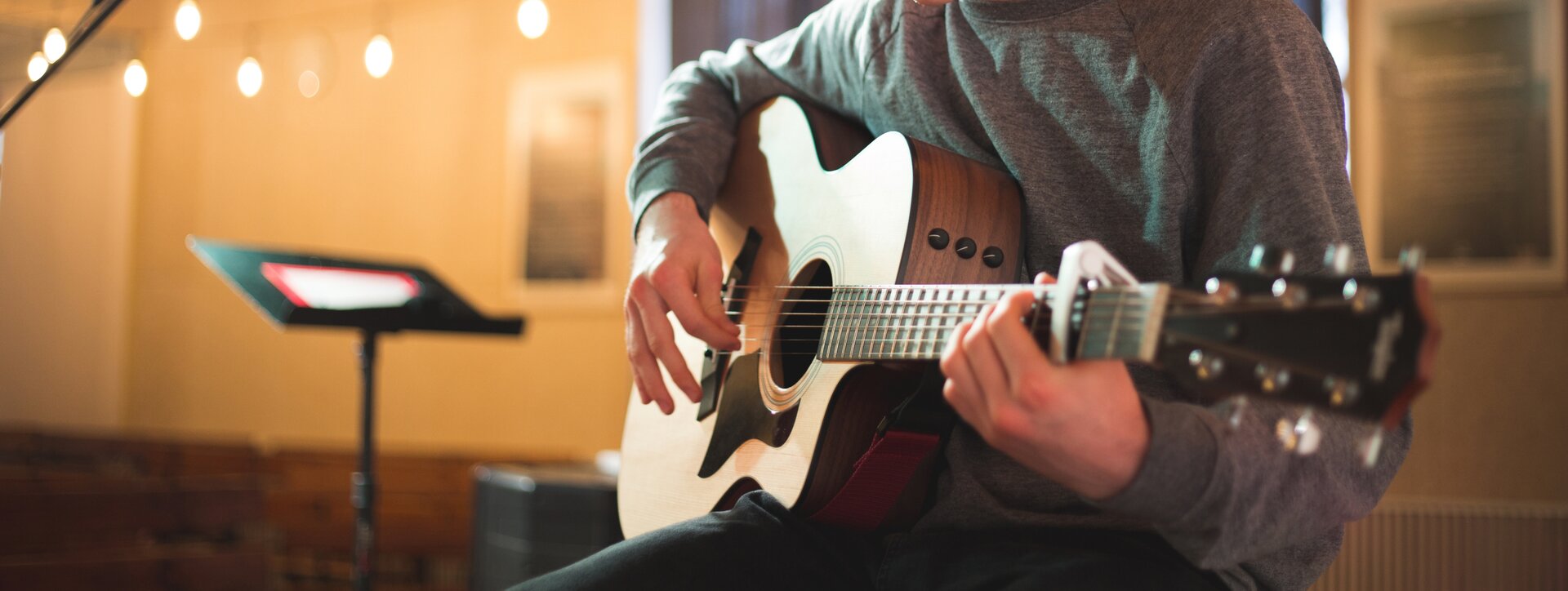 Fotografia przedstawia osobę grającą na gitarze klasycznej w pozycji siedzącej. Osoba ubrana jest w szary golf oraz ciemne spodnie. W tle pulpit wraz z partyturą utworu oraz widownia.