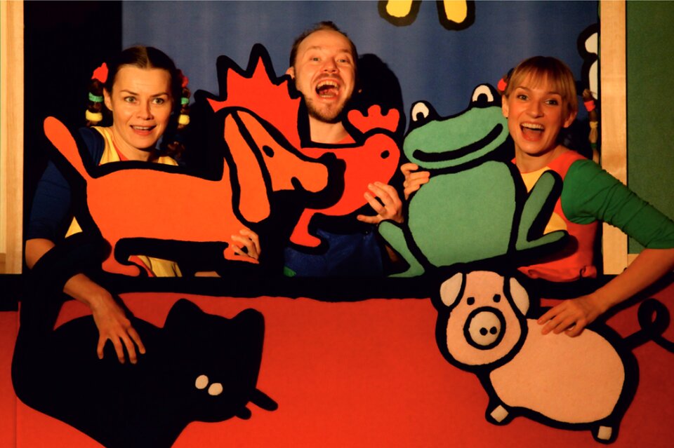 Ilustracja przedstawia zdjęcie trzech osób, trzymających w rękach rekwizyty sceniczne, przedstawiające zwierzęta. Mężczyzna trzyma czerwoną kurę, kobieta po lewej trzyma pomarańczowego psa i czarnego kota, a kobieta po prawej zieloną żabę i różową świnkę.