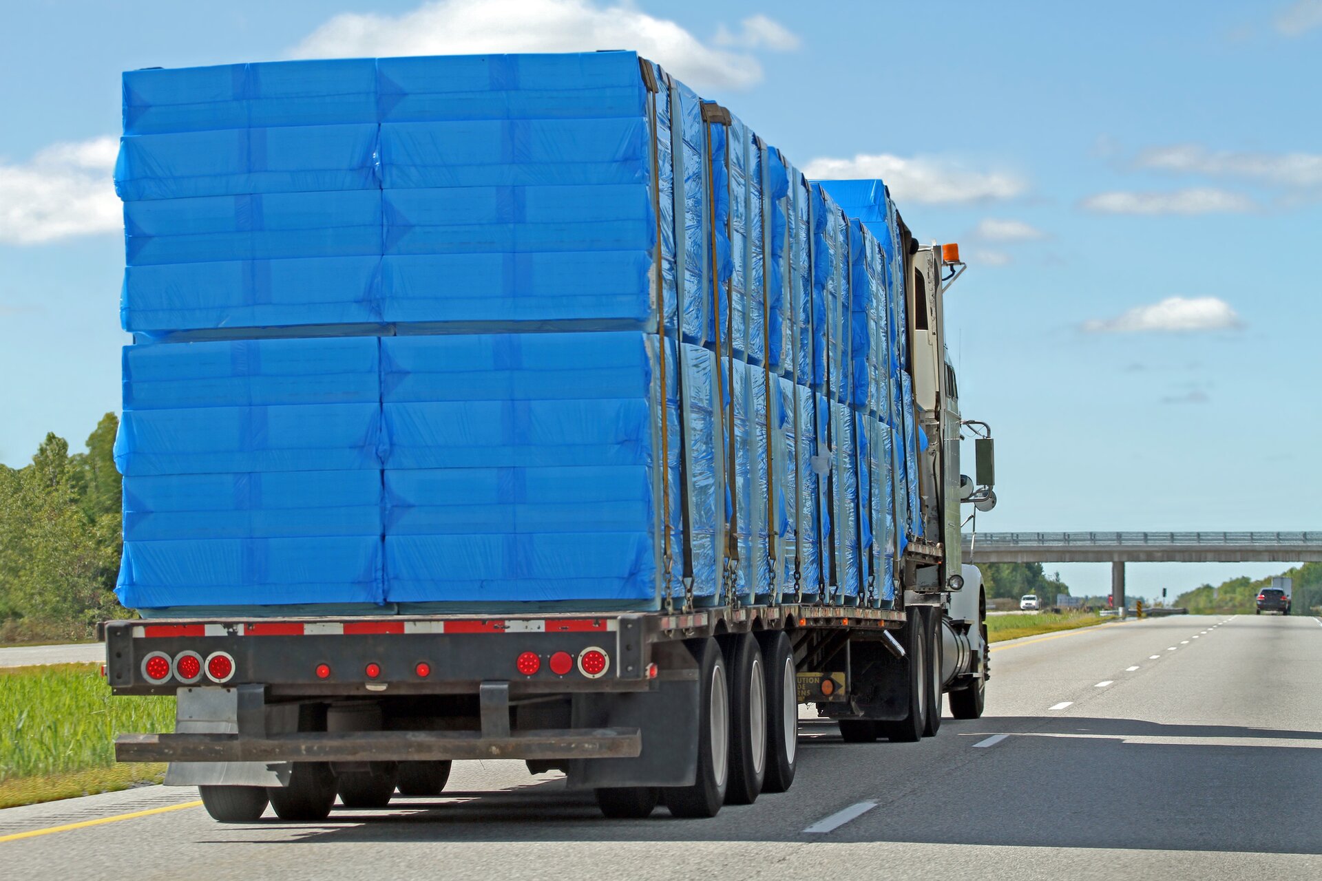 Transport styropianu.Zdjęcie przedstawia samochód ciężarowy wypełniony płasko poukładanymi płatami styropianu. Całość jest osłonięta folią dla bezpieczeństwa w transporcie.