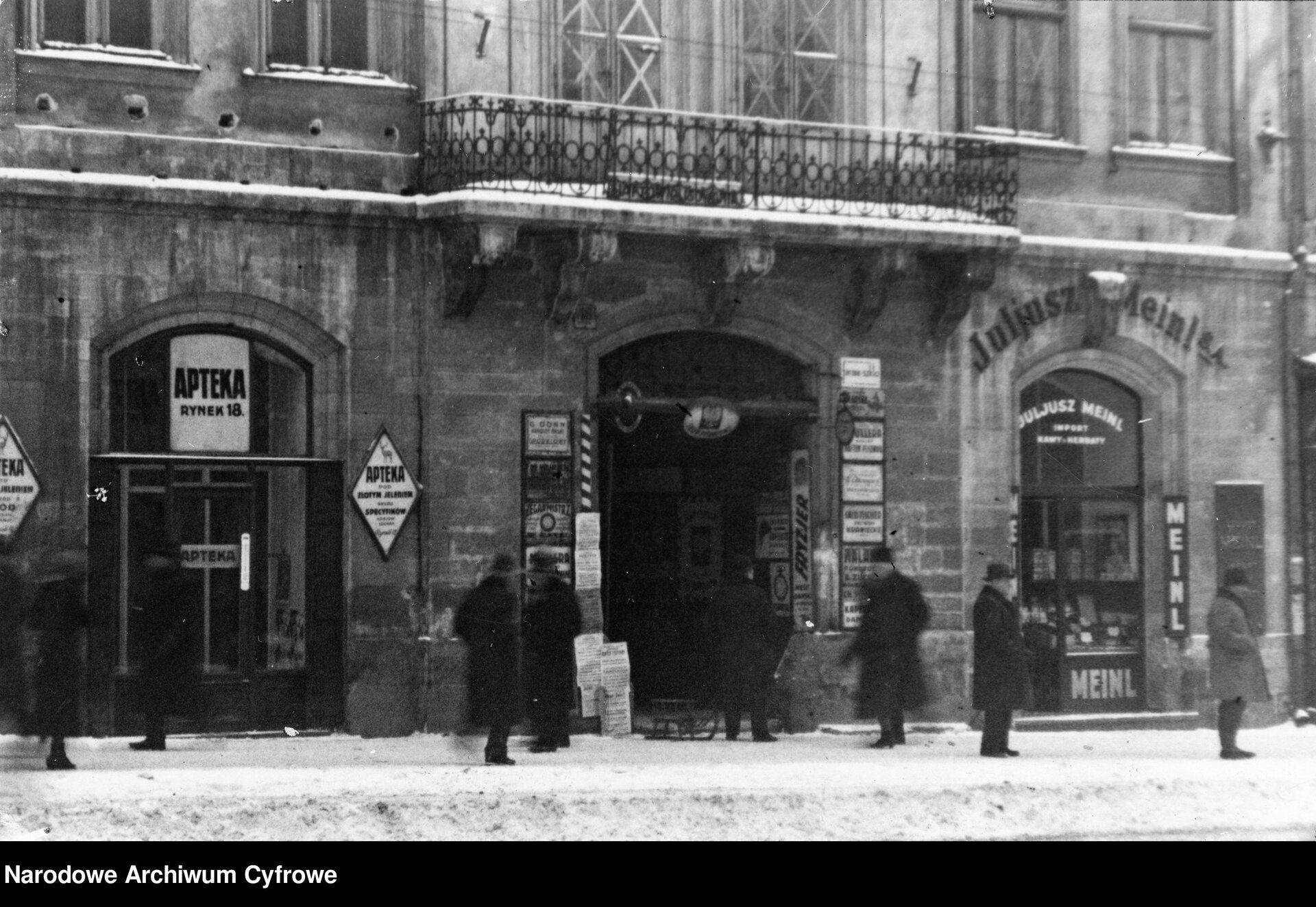Na czarno‑białej fotografii przedstawione są sklepy w Lwowie w latach trzydziestych dwudziestego wieku znajdujące się na parterze kamienicy. Obok wejścia powieszone są szyldy. Koło sklepów spacerują liczni przechodnie. Na wyższym piętrze widać balkon i okna.