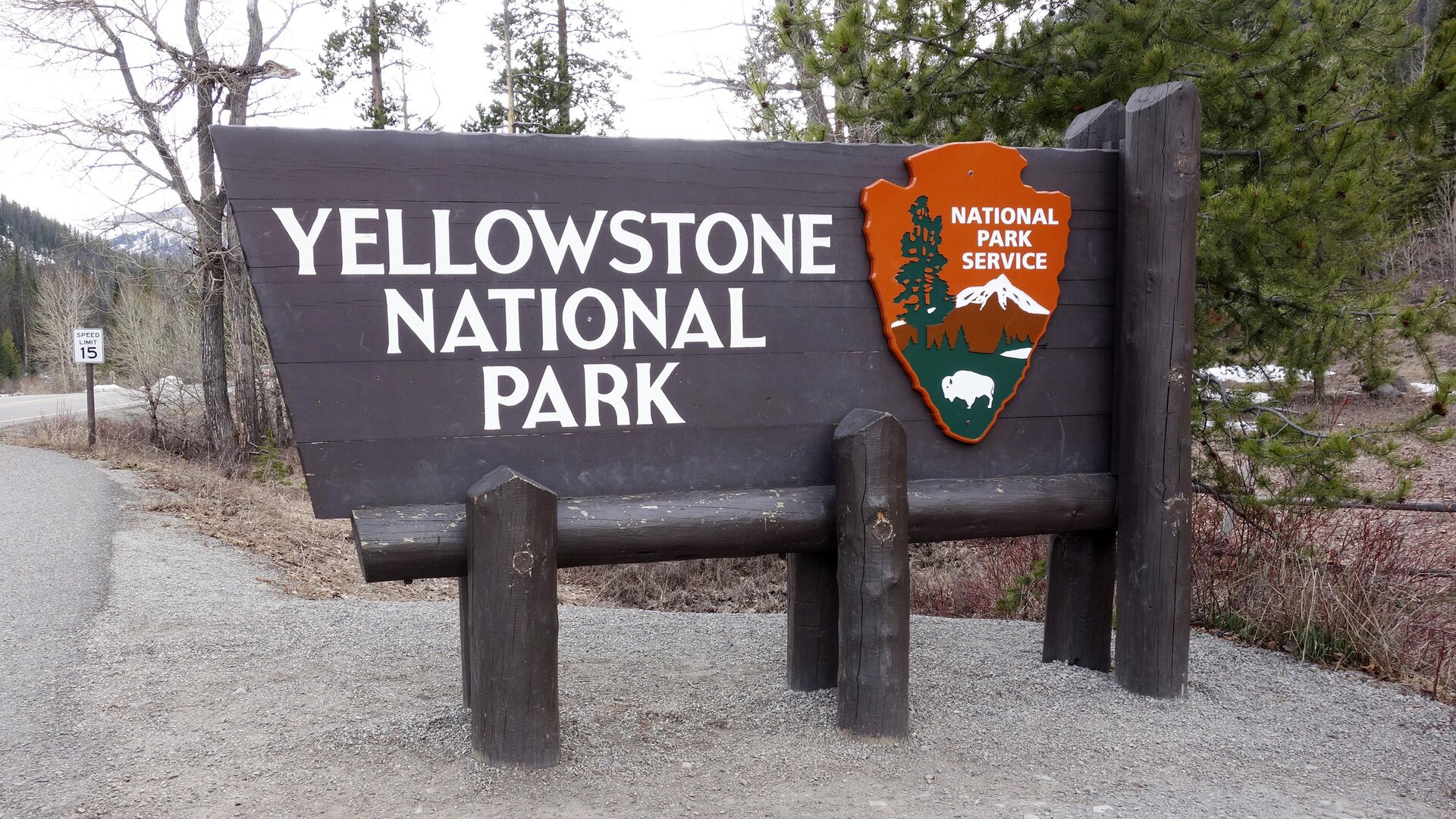Zdjęcie przedstawia tablicę informacyjną wykonaną z drewna i zamocowaną na palach. Na tablicy widnieje napis z dużych białych liter w języku angielskim: YELLOWSTONE NATIONAL PARK. Na prawo od napisu widoczny jest czerwony herb parku, na którym widoczny jest wierzchołek ośnieżonej góry, drzewo oraz bizon oraz napis z dużych białych liter w języku angielskim: NATIONAL PARK SERVICE. Znak stoi przy drodze, której fragment widoczny jest po lewej stronie zdjęcia. W tle widoczne drzewa.