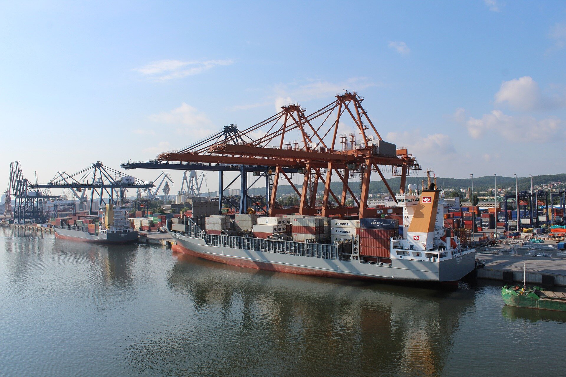 Zdjęcie kontenerowca w trakcie rozładunku. Statek jest masywny i podłużny. Znajdują się na nim dziesiątki kontenerów. Zacumowany równolegle do brzegu, nad nim stoją trzy żurawie służące do rozładunku towaru. W tle stojące na lądzie kontenery, na lewo drugi kontenerowiec.